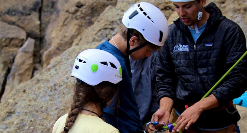 An instructor helps to tie a knot and secure it to a student's harness, while another student looks on.